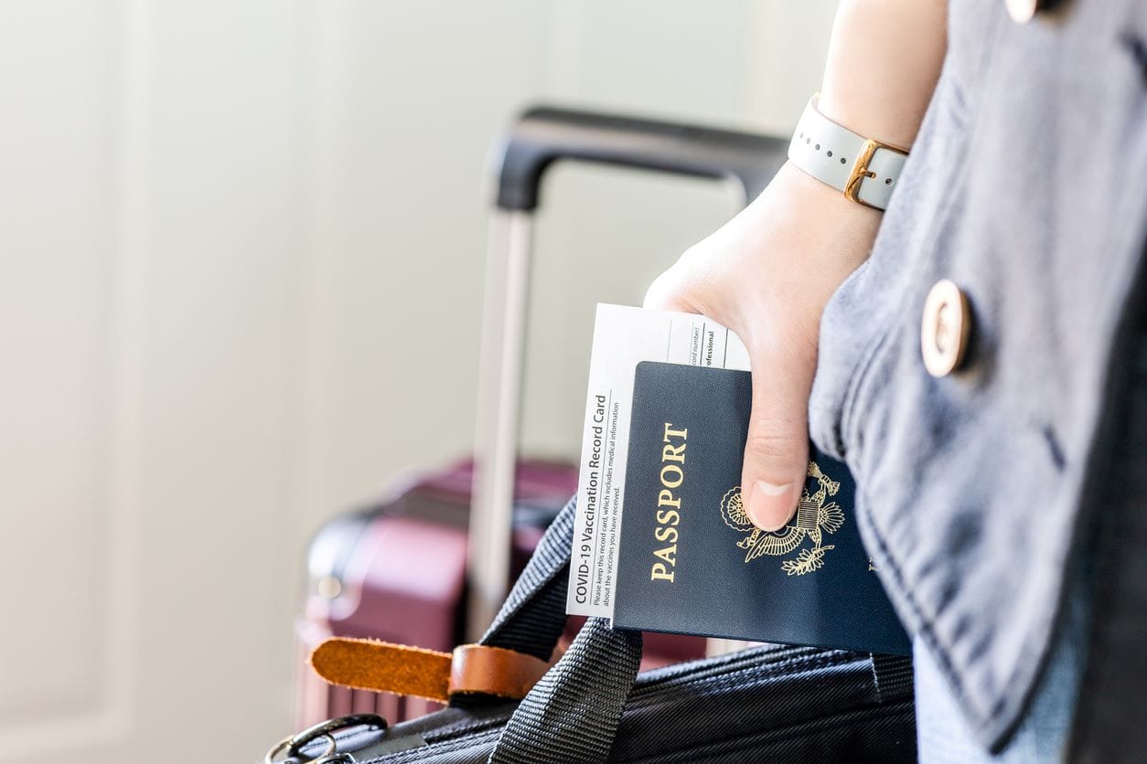 a hand grips a passport and a carryon bag with a suitcase in the background.