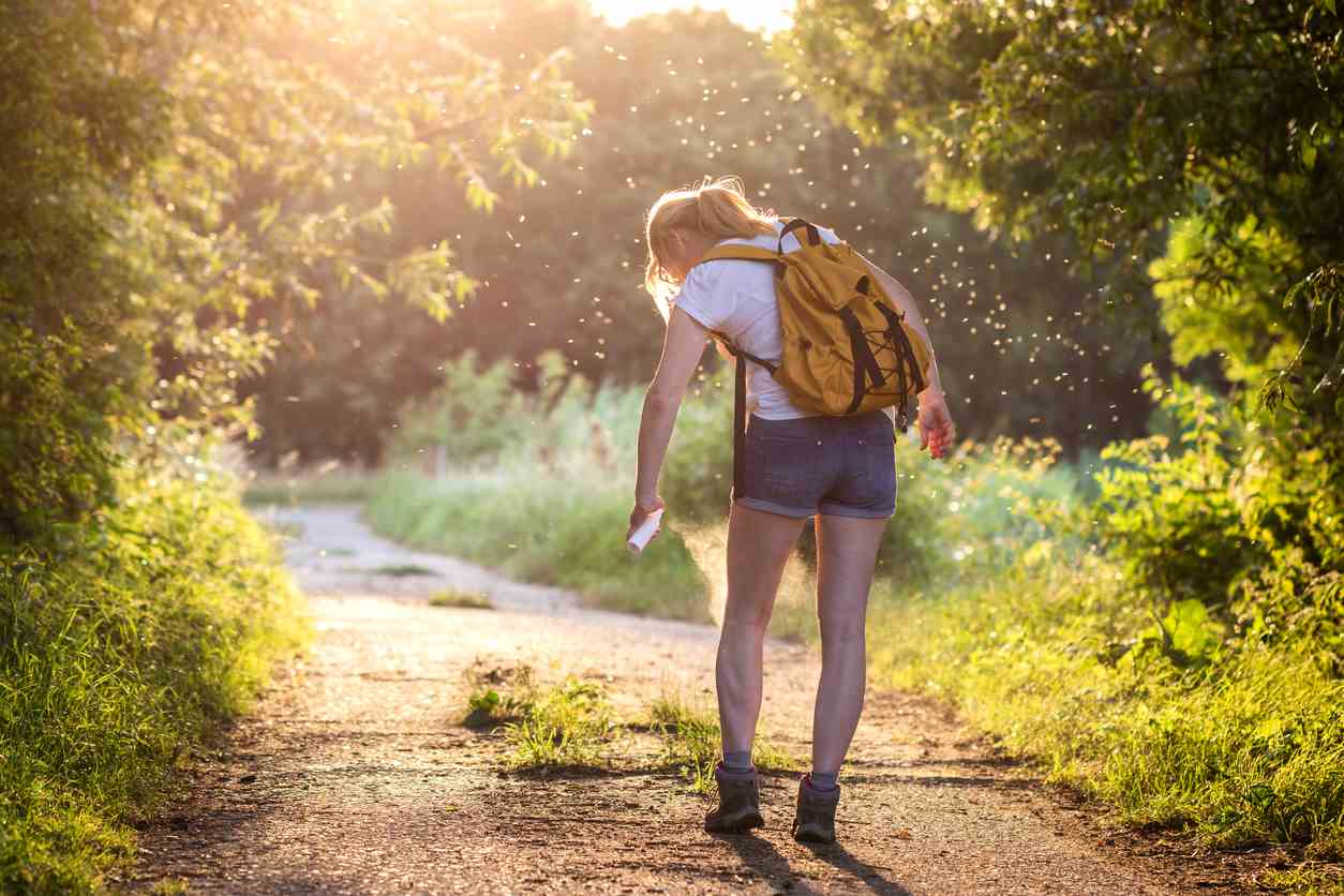 A woman applies mosquito repellent while they fly around her as she hikes on a trail.