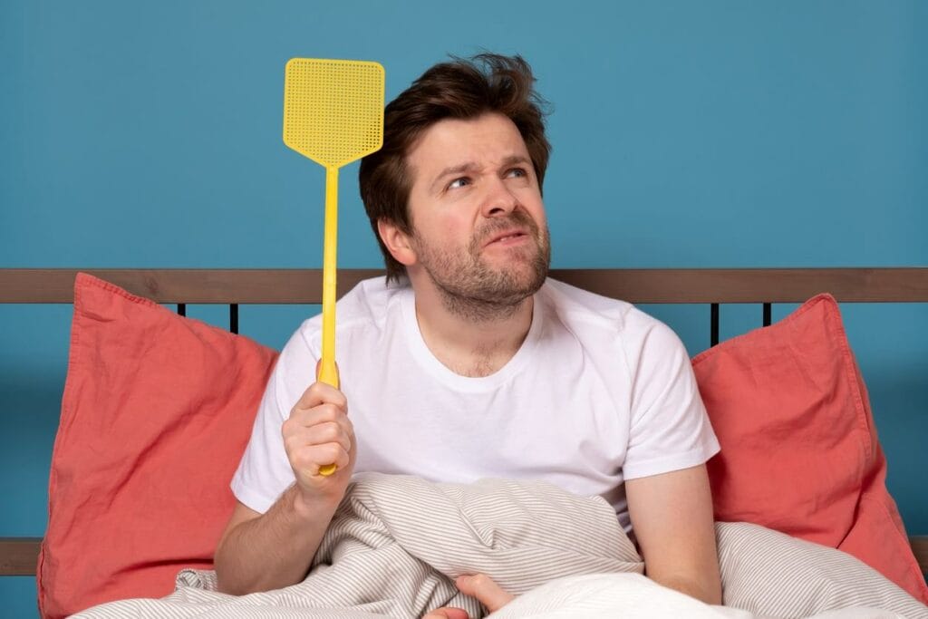 A brown-haired man in pajamas sitting up in bed, holding a yellow flyswatter while scanning the room