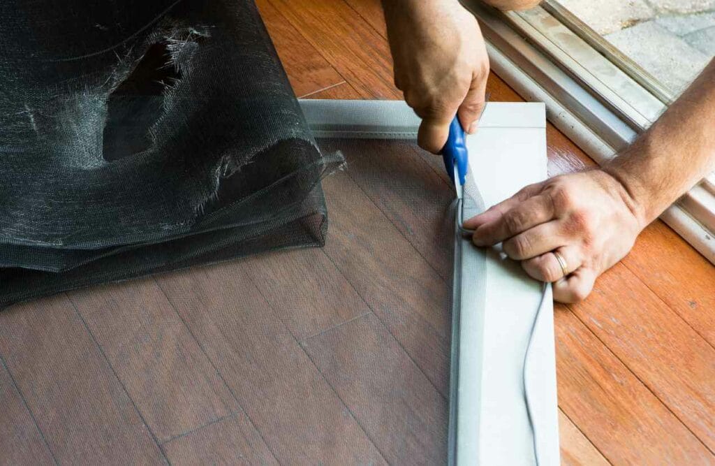 A man’s hands, wearing a wedding ring, uses a utility knife to cut a new screen for a white screen door. The old, tattered screen that’s being replaced sits in a pile just next to where he’s working.